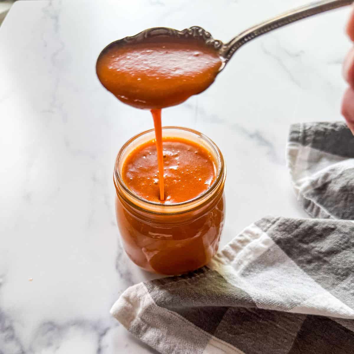 A spoon drizzles thick, reddish-orange sauce into a glass jar on a white marble surface, next to a gray and white checkered cloth.