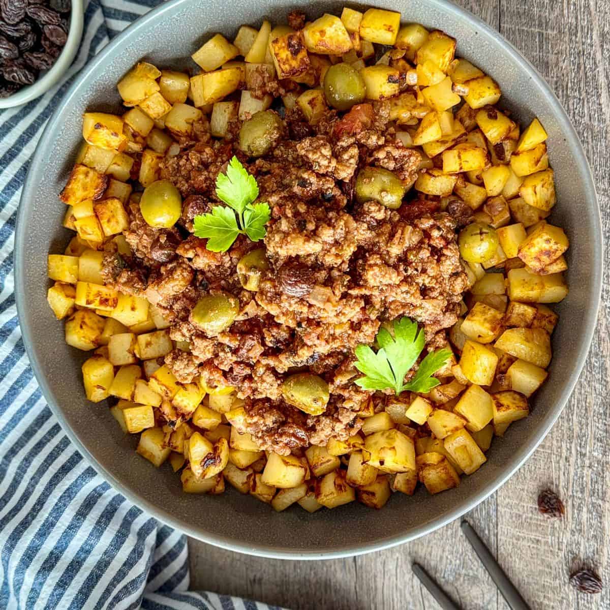 A bowl of diced, golden-brown roasted potatoes topped with ground meat, green olives, and garnished with parsley, placed on a wooden table next to a striped cloth.