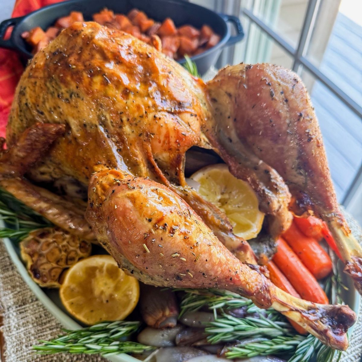 A golden roasted turkey garnished with rosemary, lemon slices, roasted garlic, carrots, and onions sits on a platter near a window, with a dish of cooked vegetables in the background.