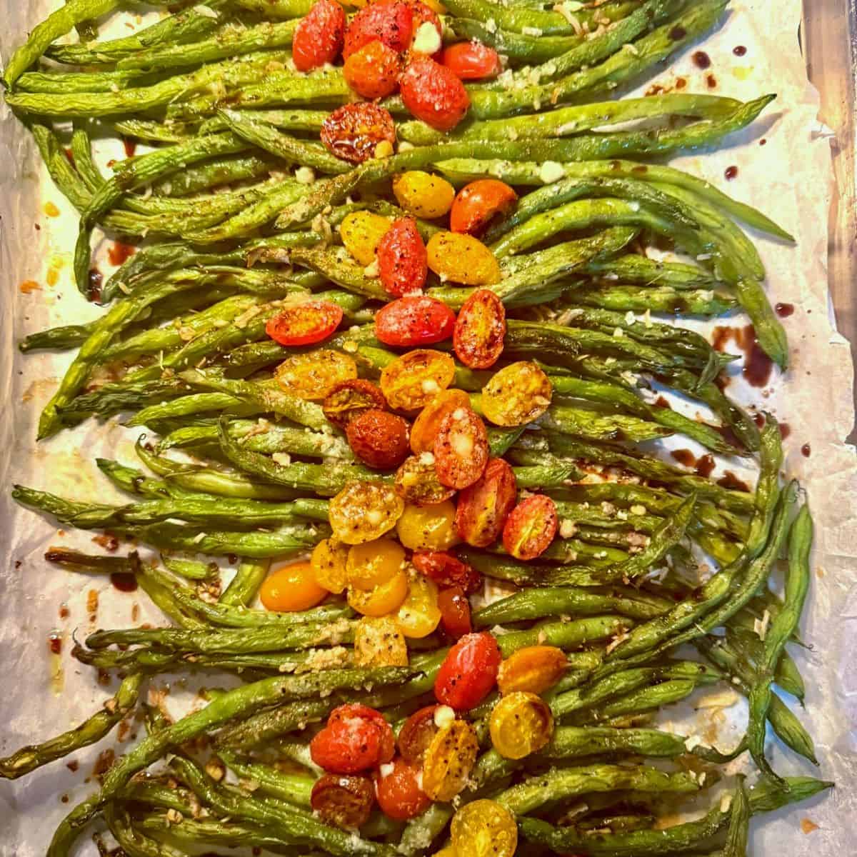 Roasted green beans topped with halved red and yellow cherry tomatoes and sprinkled with grated cheese, arranged on a parchment-lined baking sheet.