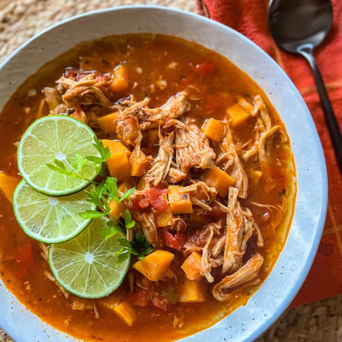A bowl of shredded chicken stew with chunks of sweet potato, diced tomatoes, and fresh herbs, garnished with three lime slices and parsley, set on a woven surface beside an orange napkin and spoon.