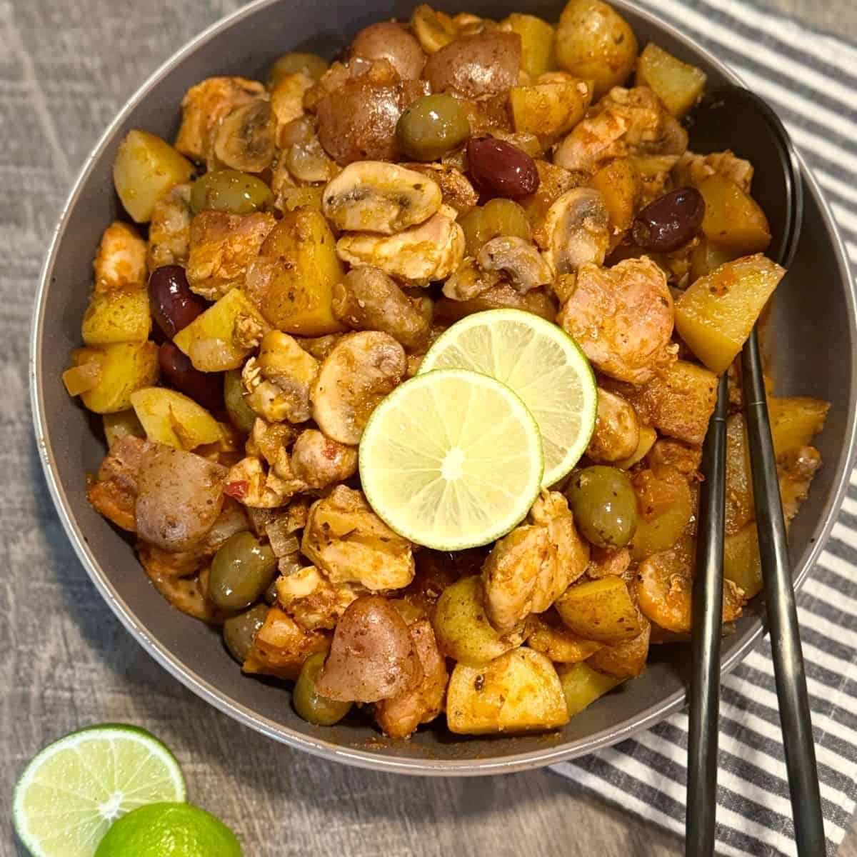 A bowl of roasted chicken, potatoes, olives, mushrooms, and cherry tomatoes, garnished with two lime slices. Black chopsticks rest on the bowl, and a sliced lime sits on a striped cloth napkin nearby.