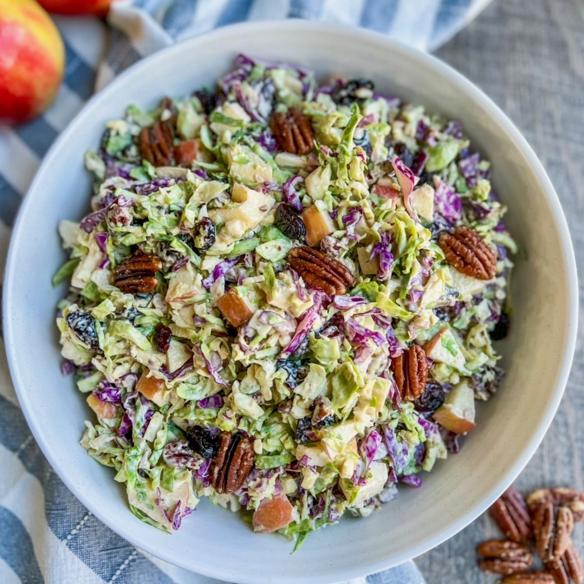 A bowl of colorful coleslaw with chopped green and purple cabbage, diced apples, pecans, and raisins, all mixed with a creamy dressing. The bowl is placed on a blue and white striped cloth.