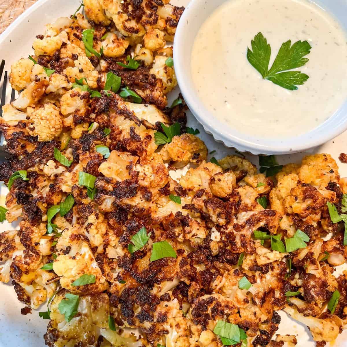 A plate of roasted cauliflower garnished with chopped herbs, served with a bowl of creamy white dipping sauce topped with a parsley leaf.