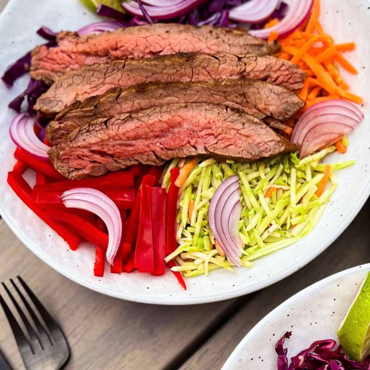 A bowl filled with sliced grilled steak, shredded carrots, red cabbage, broccoli slaw, red bell peppers, and red onion slices, arranged in colorful sections. A fork and knife are visible nearby on a wooden surface.