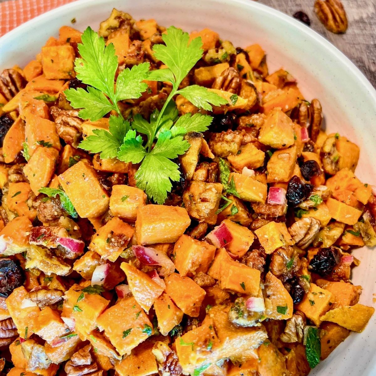 A close-up of a bowl filled with diced sweet potato salad, garnished with fresh parsley. The salad contains red onions, pecans, and dried cranberries, all mixed together in a light dressing.