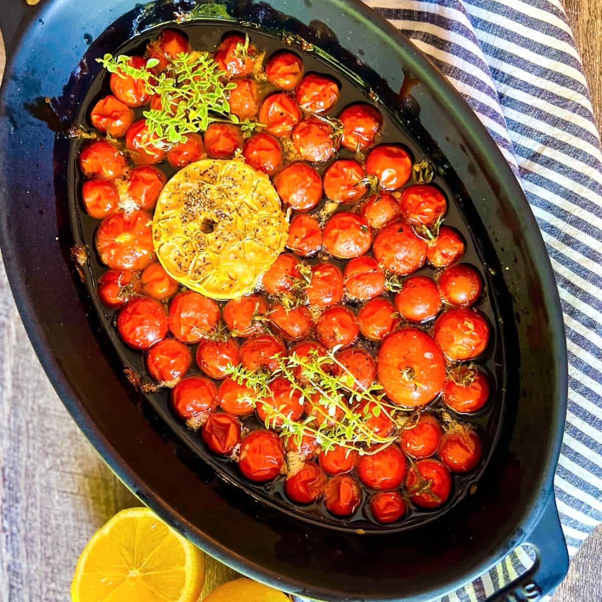 An oval black baking dish holds roasted cherry tomatoes, a halved head of garlic, sprigs of fresh thyme, and a drizzle of olive oil. A striped towel and halved lemon are placed beside the dish on a wooden surface.