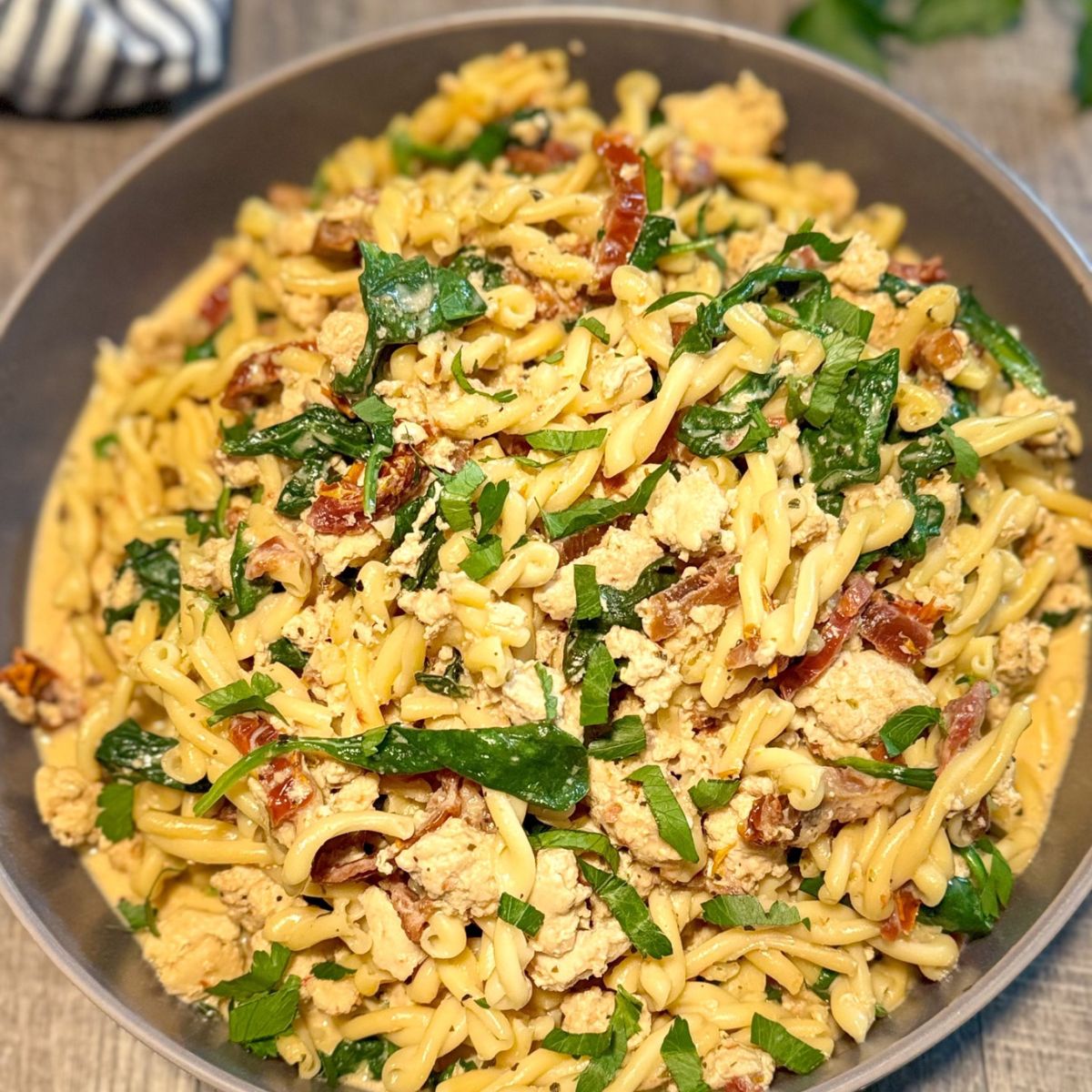 A bowl of pasta mixed with spinach, sun-dried tomatoes, crumbled tofu, and fresh herbs, served on a wooden surface.