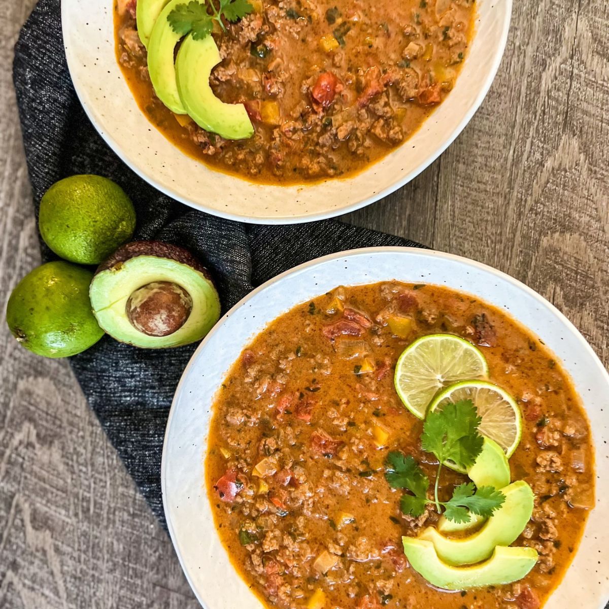 Two bowls of creamy taco soup topped with sliced avocado, lime, and cilantro. Next to the bowls are whole and halved avocados and fresh limes, arranged on a dark cloth on a wooden surface.