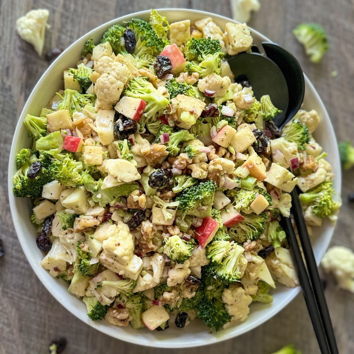 A bowl of broccoli salad with chopped apples, cauliflower, raisins, walnuts, and red onions, mixed with a creamy dressing. A serving spoon and fork are placed in the bowl, with some ingredients scattered on the wooden table.