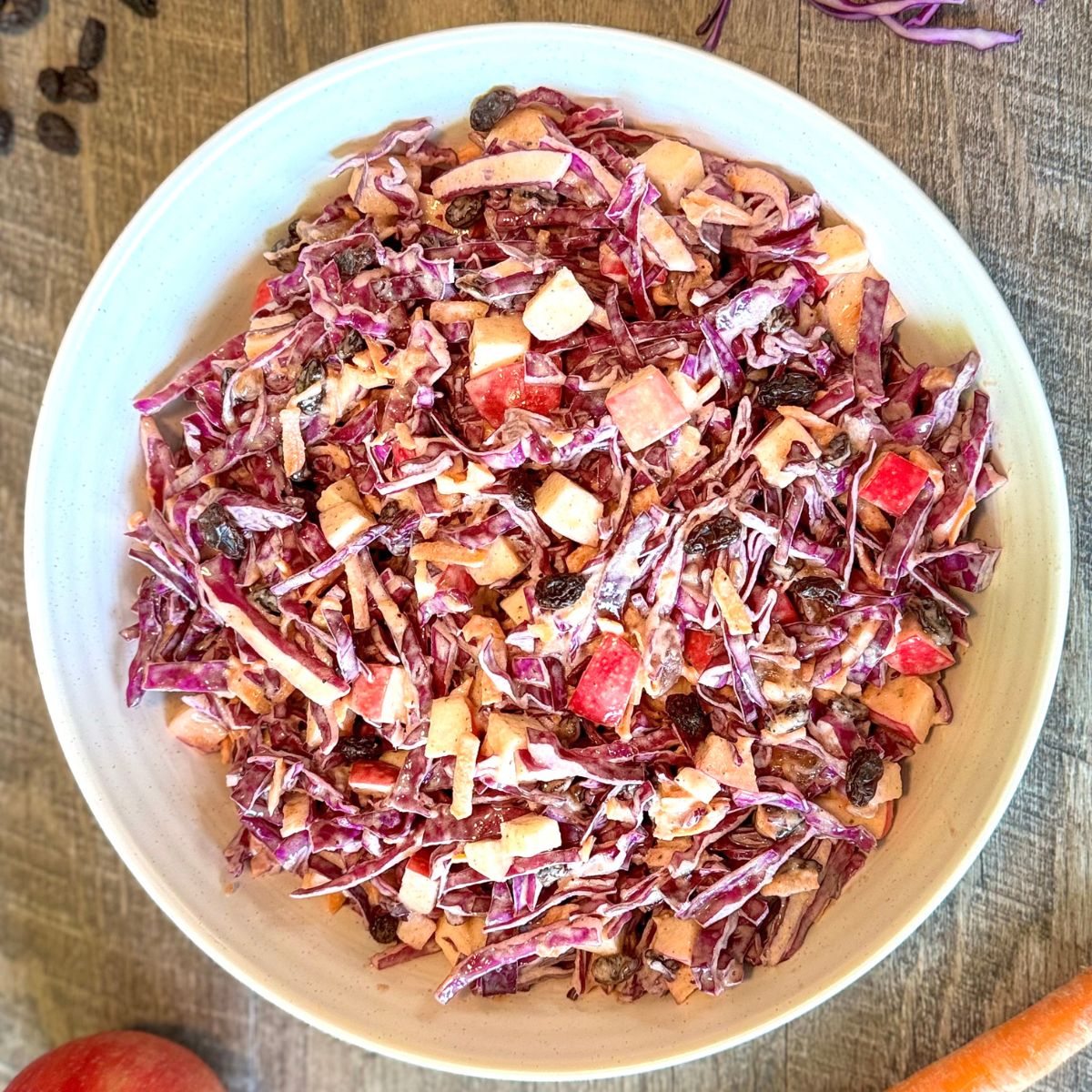 A bowl filled with colorful coleslaw made of shredded red cabbage, diced apples, raisins, and carrots, all coated in a creamy dressing, placed on a wooden surface.