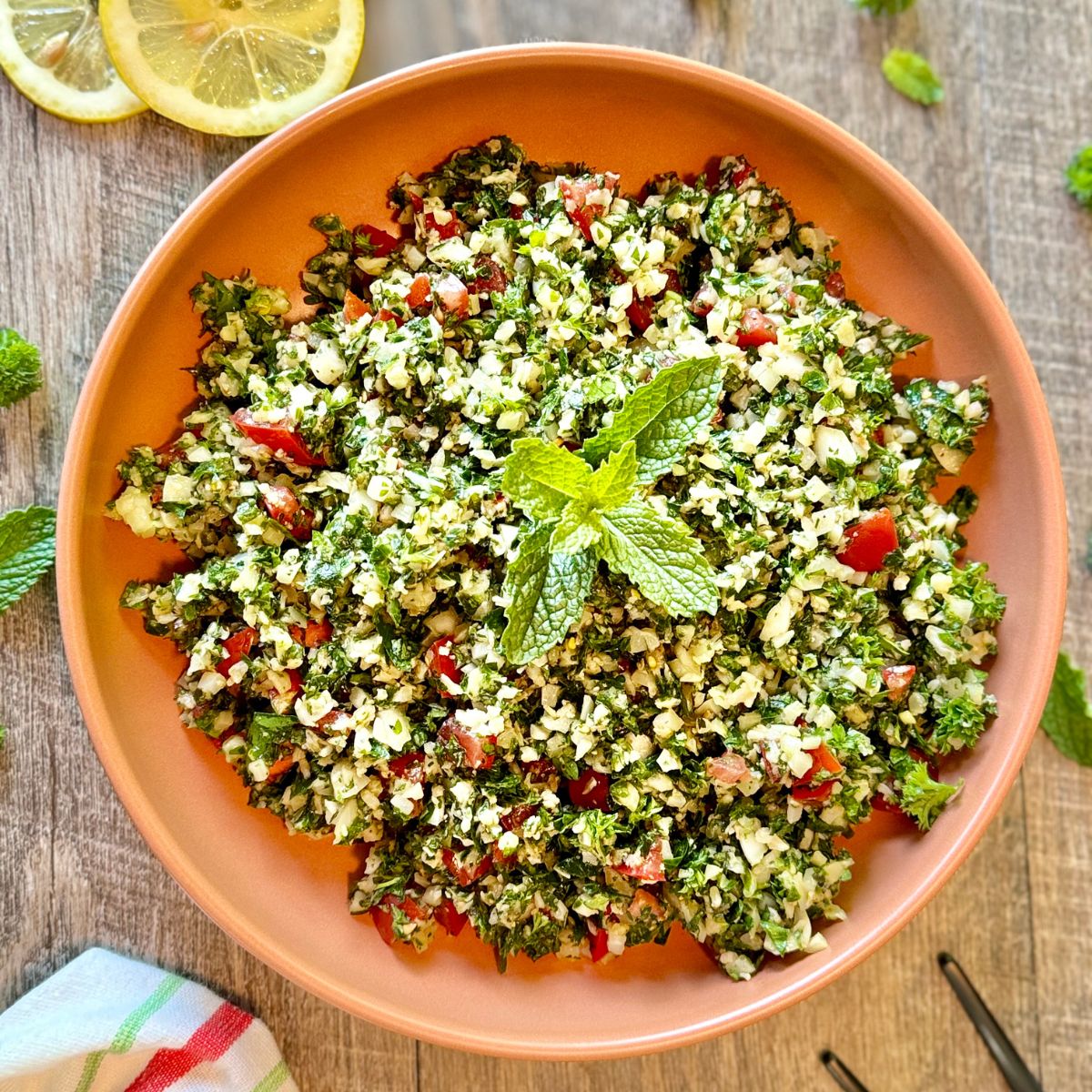 A bowl of tabbouleh salad topped with a fresh mint sprig, featuring finely chopped parsley, tomatoes, and cauliflower, placed on a wooden surface with lemon slices and mint leaves nearby.