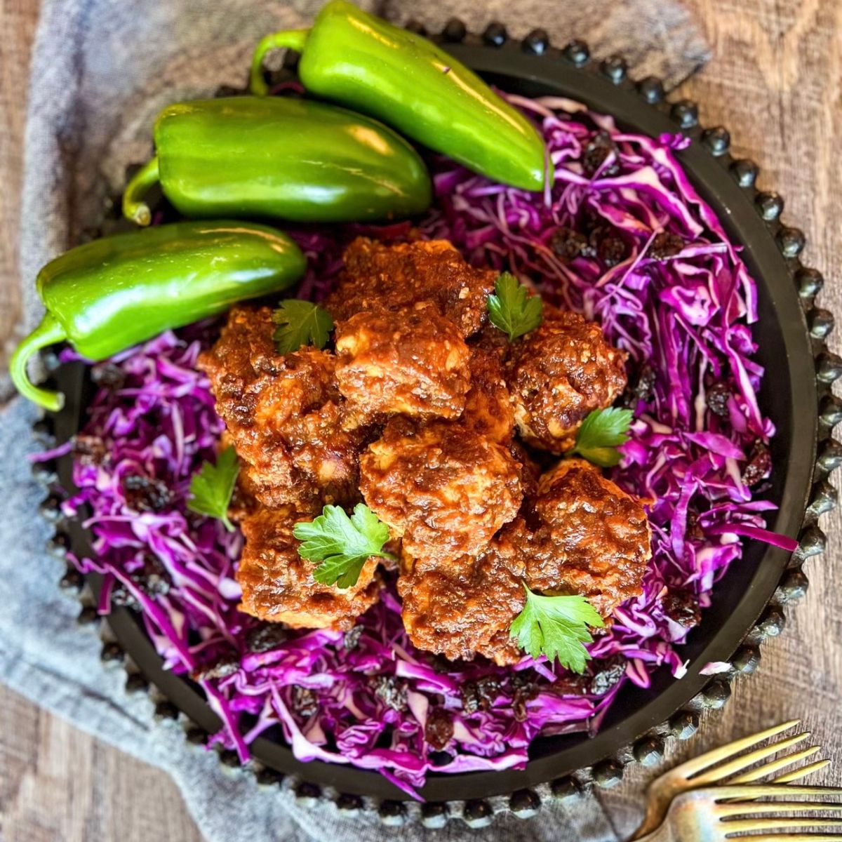 A black plate with spicy meatballs on shredded purple cabbage, garnished with parsley, and three whole green jalapeños on the side; a fork and gray napkin nearby on a wooden surface.