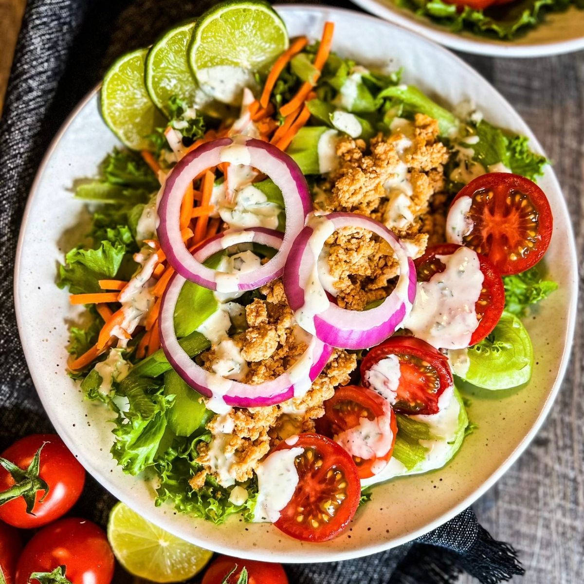 A fresh salad in a bowl topped with crispy breaded pieces, sliced red onion, cherry tomatoes, shredded carrots, lime wedges, greens, and drizzled ranch dressing, with whole cherry tomatoes beside the bowl.