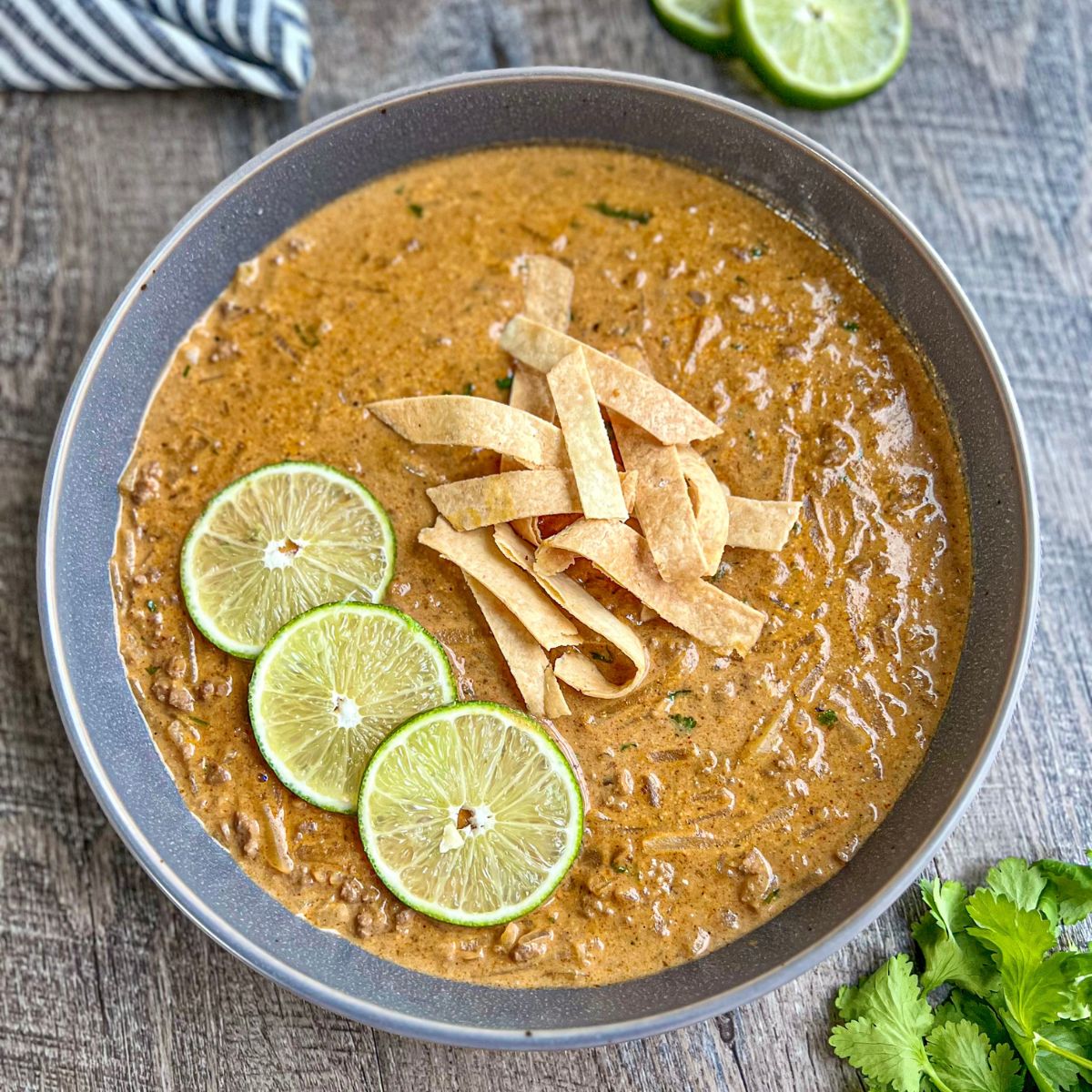 A bowl of creamy soup garnished with three lime slices and crispy tortilla strips, set on a wooden table with a sprig of cilantro and lime wedges nearby.