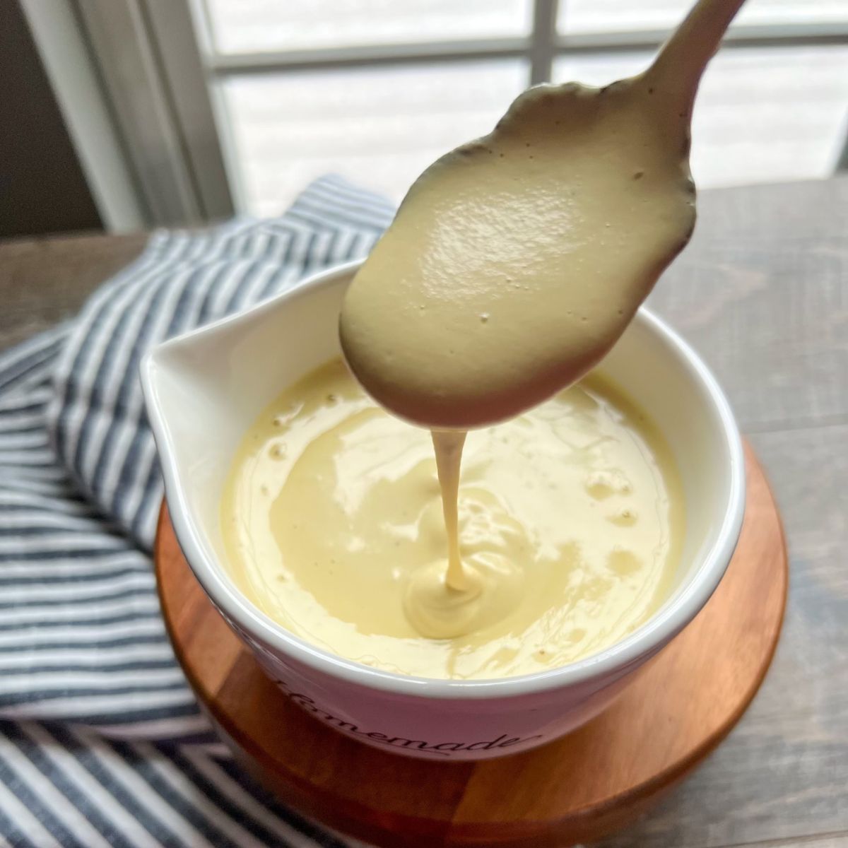 A spoon drizzling thick, creamy yellow sauce into a white bowl set on a wooden surface, with a striped cloth nearby and a window in the background.