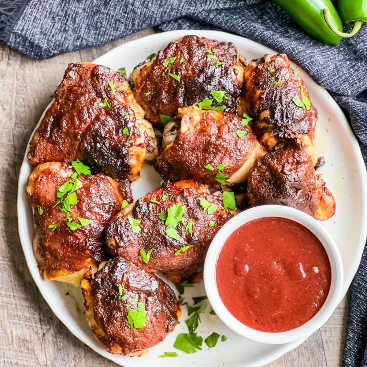 A plate of barbecued chicken thighs garnished with chopped parsley, served with a small bowl of red barbecue sauce on the side, placed on a wooden surface with a dark cloth and a green chili pepper nearby.