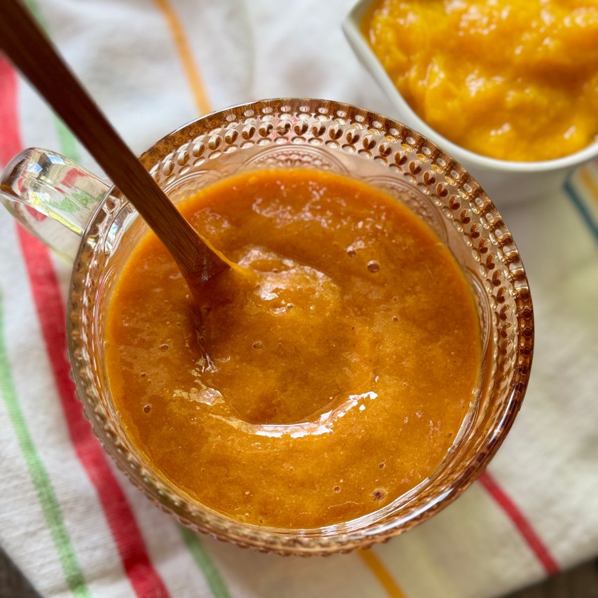 A glass bowl filled with thick orange sauce or puree, being stirred with a wooden spoon, sits on a striped cloth. In the background, there is a small white bowl containing more of the same puree.