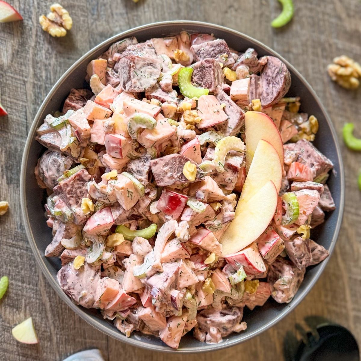 A bowl of salad with diced apples, celery, walnuts, and chunks of beets, all mixed in a creamy dressing, garnished with apple slices. The bowl is set on a wooden surface with scattered salad ingredients.