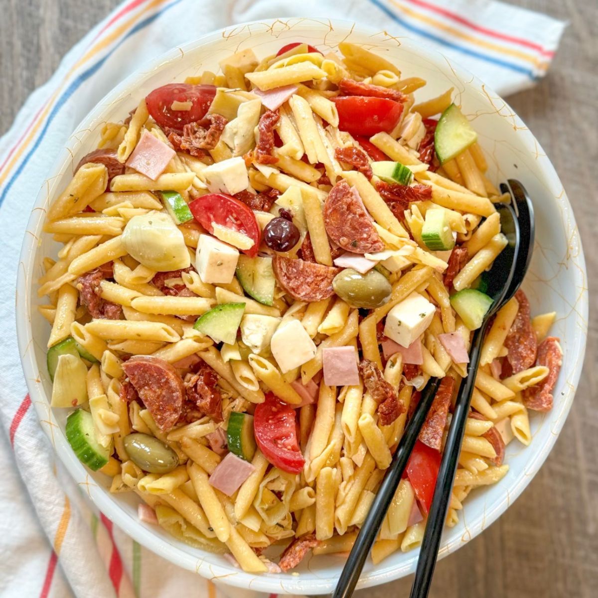 A bowl of pasta salad with penne, cherry tomatoes, cucumber, olives, cheese cubes, pepperoni, ham, artichoke hearts, and green onions, served with black utensils on a striped napkin.