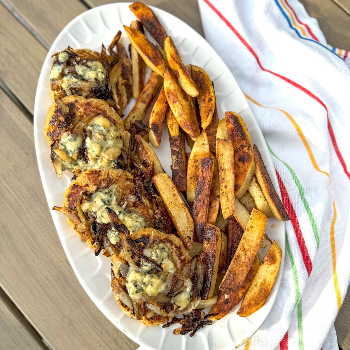 A white oval plate holds roasted potato wedges and three beef patties topped with melted blue cheese and caramelized onions. A striped kitchen towel lies beside the plate on a wooden table.