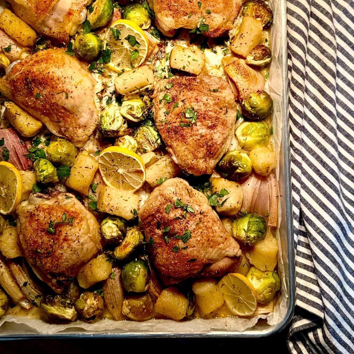 A sheet pan filled with roasted chicken thighs, Brussels sprouts, potatoes, shallots, and lemon slices, garnished with fresh herbs. A striped towel is visible beside the pan.