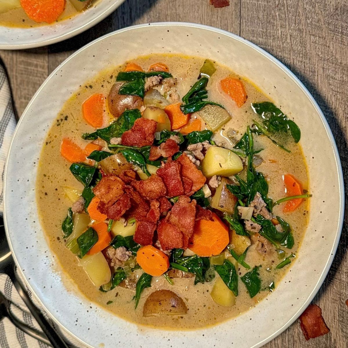 A bowl of creamy soup filled with sliced carrots, potatoes, spinach, ground meat, and topped with crispy bacon pieces, set on a wooden table.