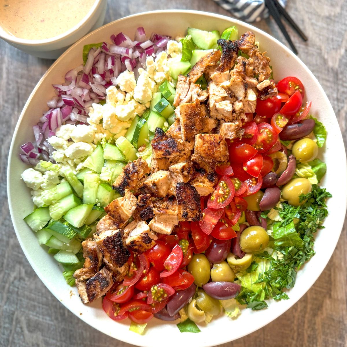 A colorful Greek salad in a bowl topped with grilled chicken, cherry tomatoes, olives, cucumbers, red onions, feta cheese, and fresh herbs, arranged in neat rows. A small bowl of dressing is visible in the corner.