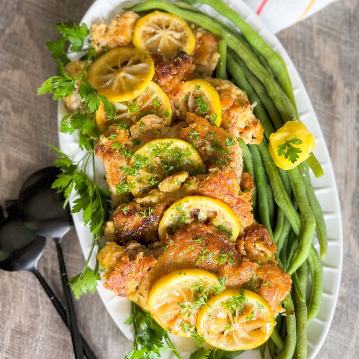 A platter of golden-brown, breaded chicken thighs topped with lemon slices and fresh herbs, garnished with long green beans and parsley, set on a wooden table. Black serving utensils are beside the dish.