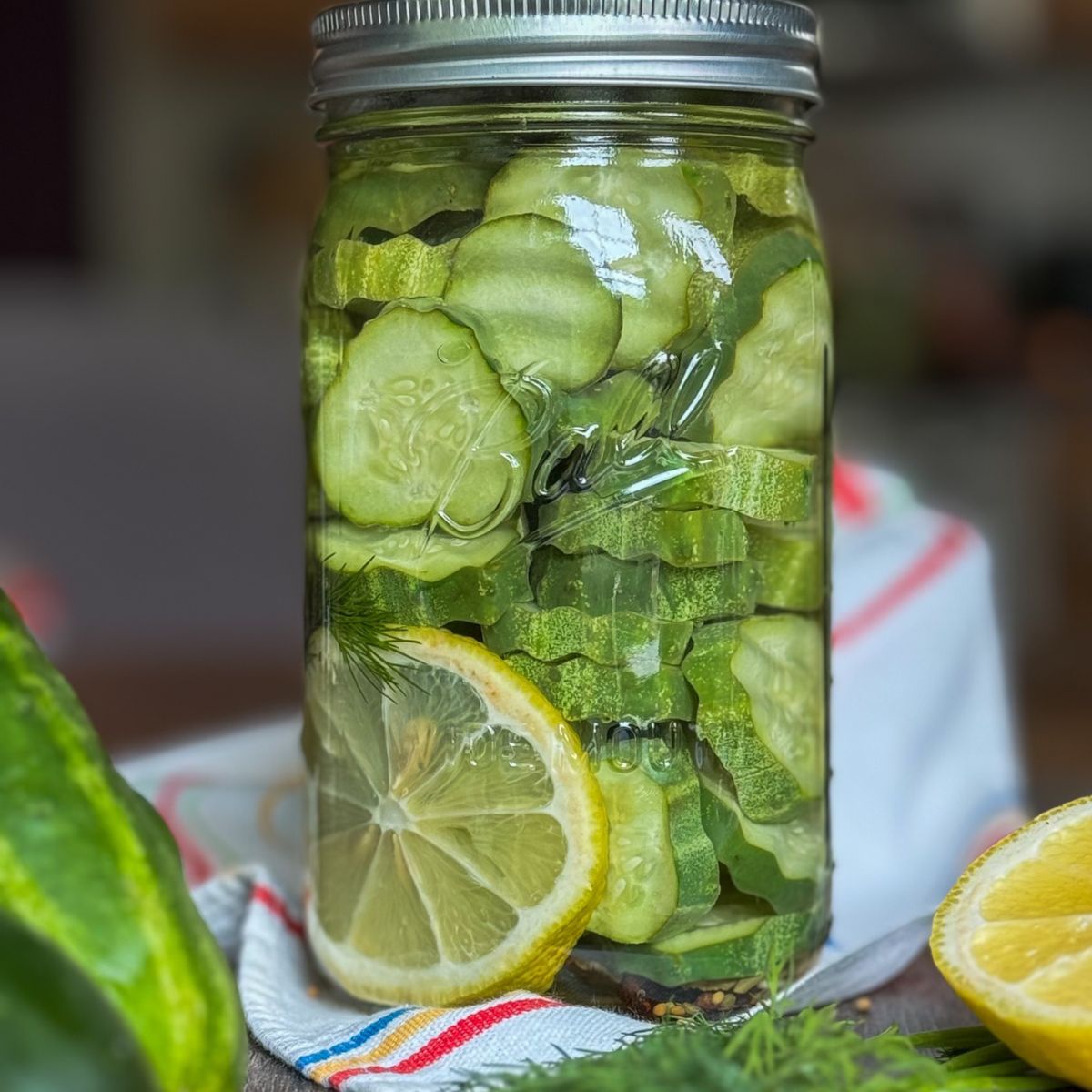A mason jar filled with sliced cucumbers, sitting on a cloth with lemon slices, dill, and peppercorns nearby. The jar is tightly sealed and surrounded by fresh ingredients.