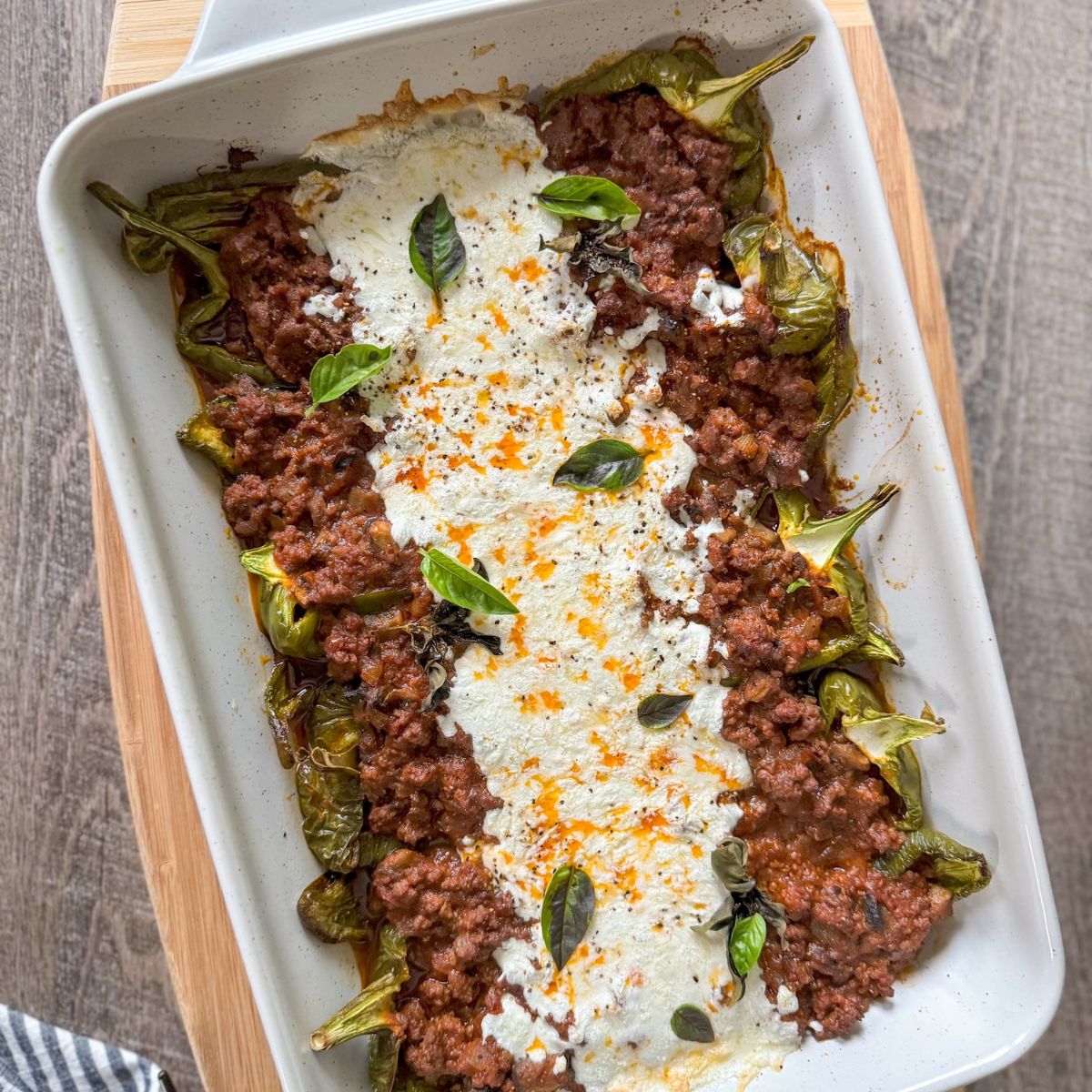 A white baking dish filled with stuffed green peppers, topped with melted cheese, tomato meat sauce, fresh basil leaves, and sprinkled seasoning, placed on a wooden surface.
