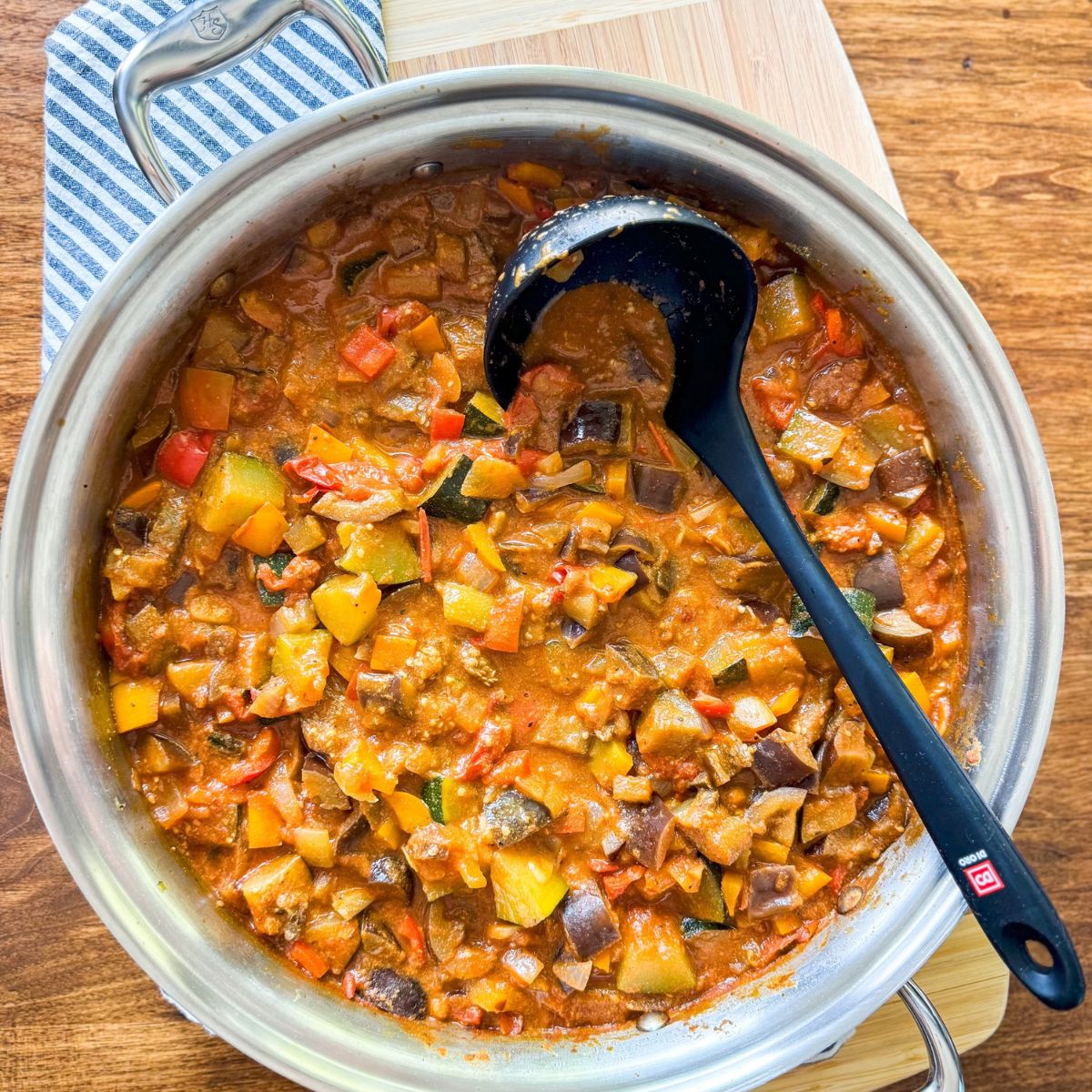 A stainless steel pan filled with colorful ratatouille made of diced vegetables in a tomato-based sauce, with a black ladle resting in the pan, on a wooden table with a striped cloth nearby.