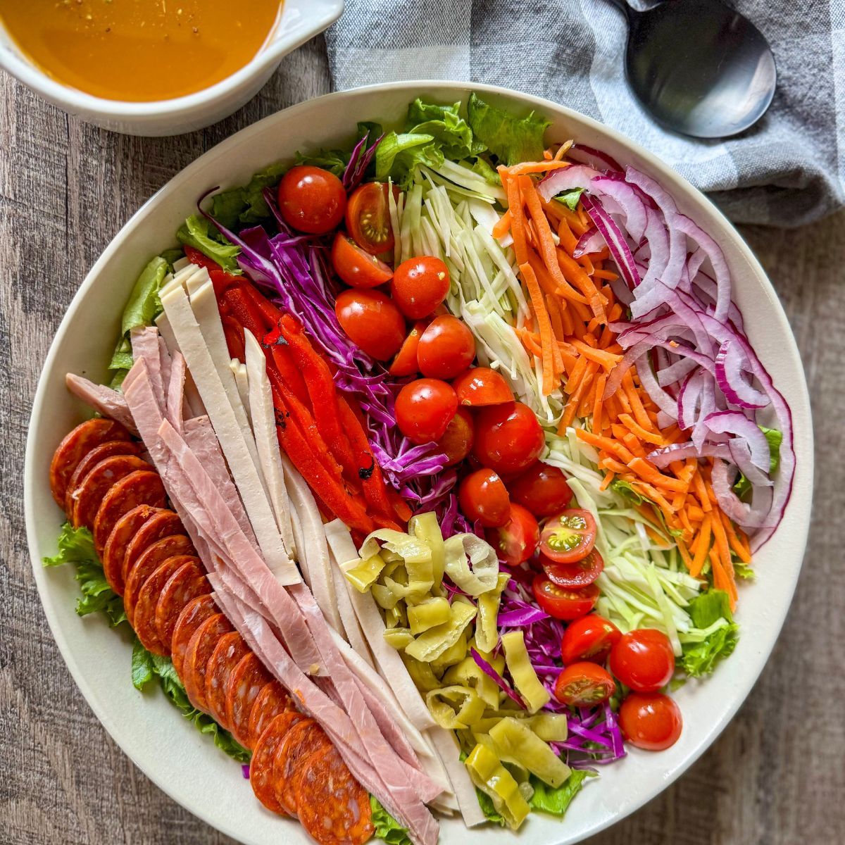 A large bowl filled with a colorful salad, featuring rows of sliced pepperoni, ham, red peppers, shredded cabbage, cherry tomatoes, carrots, onions, and green olives, with a bowl of dressing and spoon nearby.