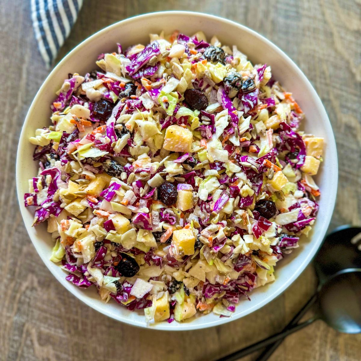 A bowl of colorful coleslaw with shredded cabbage, carrots, raisins, and diced apples sits on a wooden table next to black serving utensils.