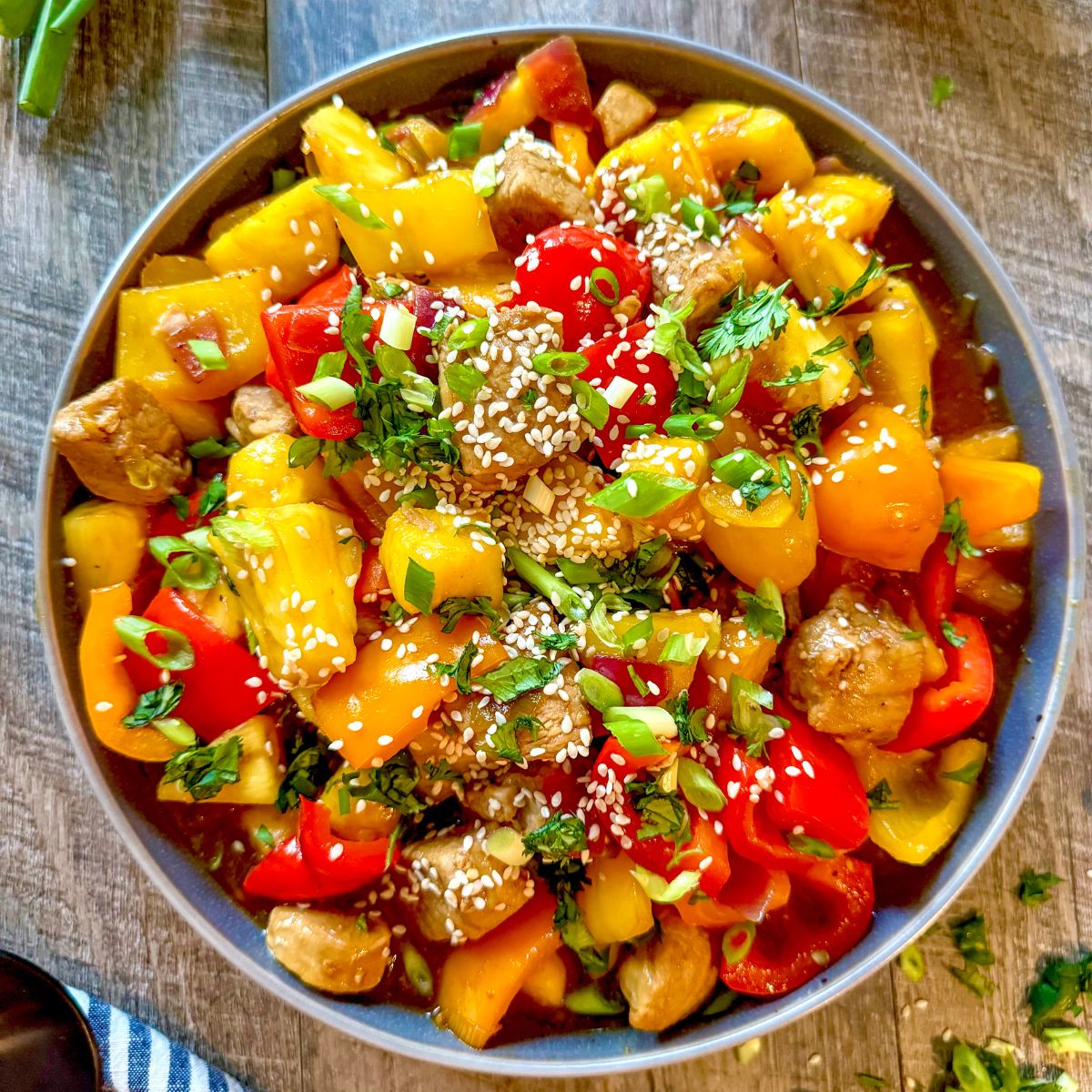 A colorful bowl of sweet and sour pork with chunks of pineapple, red and yellow bell peppers, cherry tomatoes, green onions, fresh herbs, and sprinkled with sesame seeds on a wooden table.
