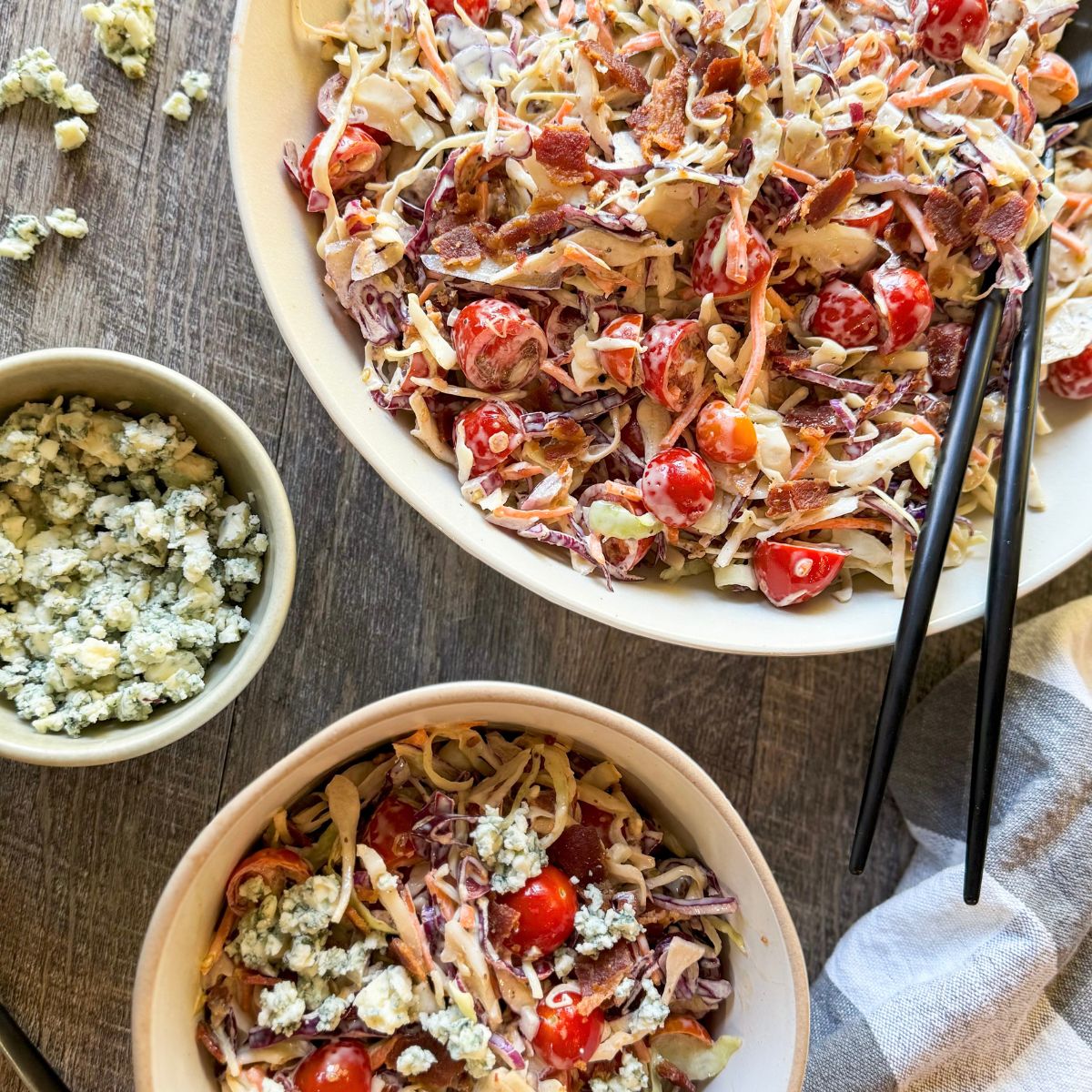Two bowls of salad with shredded cabbage, cherry tomatoes, and bacon bits sit on a wooden table. A small bowl of crumbled blue cheese is nearby. Chopsticks and a striped towel are beside the bowls.