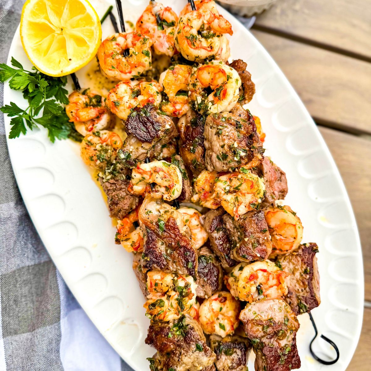 Skewers of grilled shrimp and steak are arranged on a white oval platter, garnished with fresh herbs and a halved lemon, next to a gray and white cloth on a wooden table.