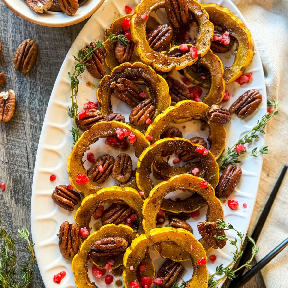 A white oval platter with roasted delicata squash rings, pecans, and pomegranate seeds, garnished with fresh thyme sprigs on a rustic wooden surface.