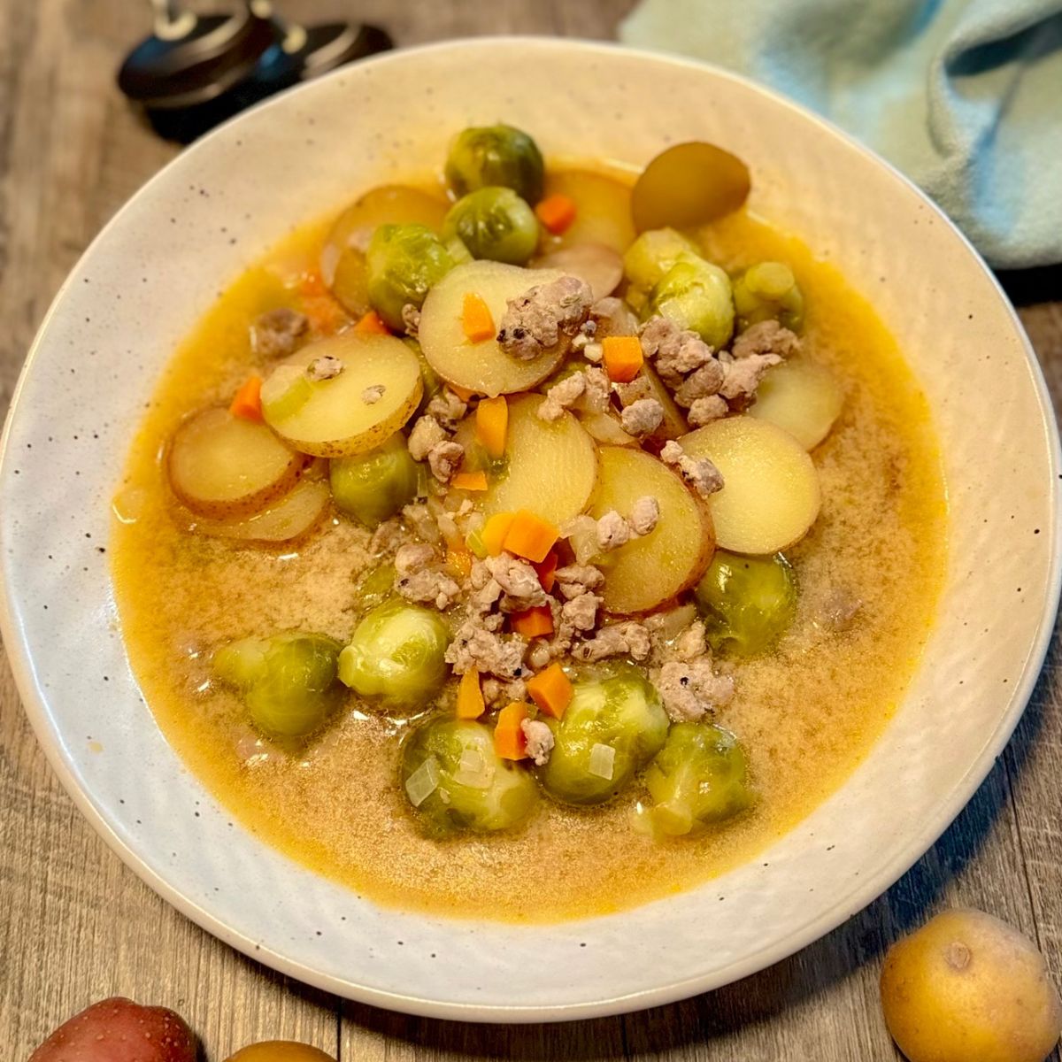 A bowl of soup with sliced potatoes, Brussels sprouts, ground meat, and diced carrots in a light broth, served on a wooden table with a blue cloth and scattered potatoes nearby.