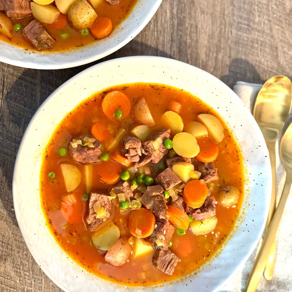 Two white bowls filled with beef stew containing chunks of beef, potatoes, carrots, and peas in a rich broth, placed on a wooden surface with gold utensils to the side.