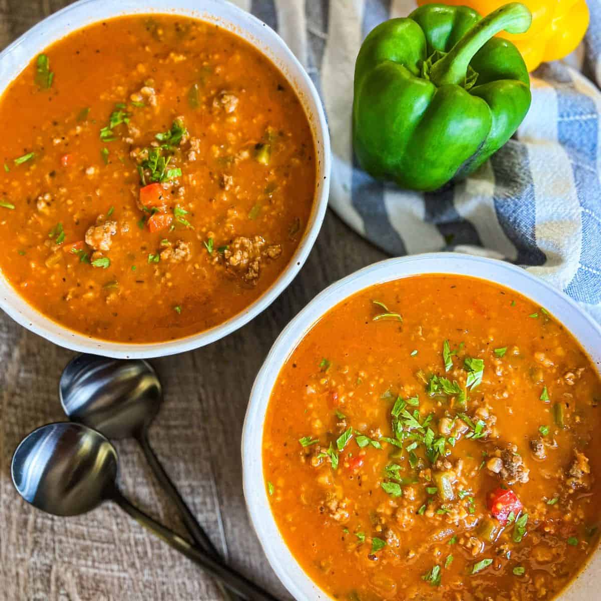 Two bowls of hearty soup with ground meat, tomatoes, and herbs on a wooden table. A green bell pepper and a striped cloth are nearby, with two metal spoons placed beside the bowls.
