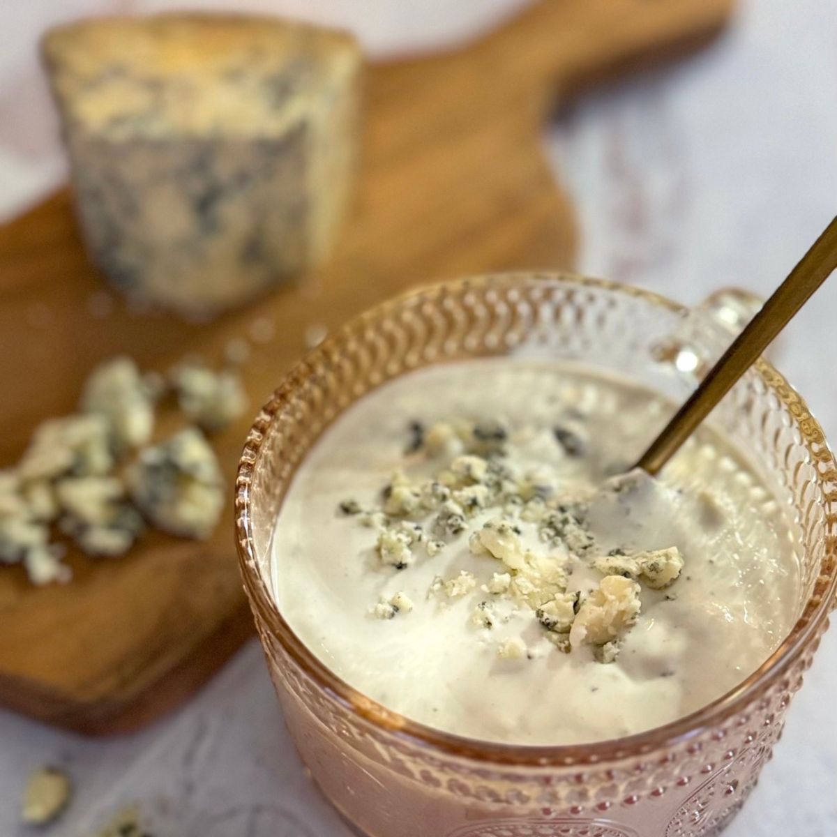 A glass cup filled with creamy dip topped with crumbled blue cheese, with a spoon inside. In the background, a wooden board holds more crumbled blue cheese and a cheese wedge.