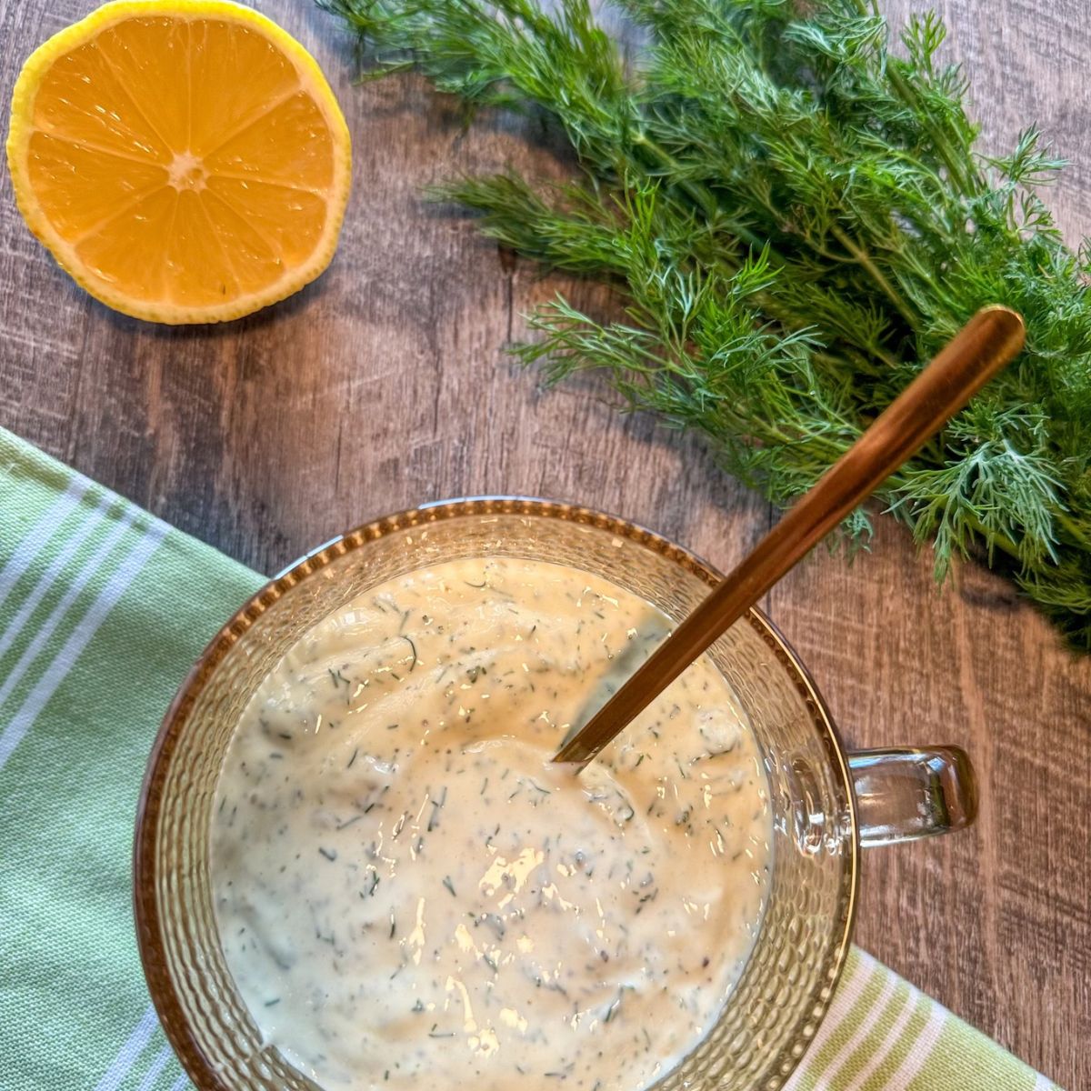 A glass bowl of creamy dill sauce with a spoon inside, placed on a wooden surface next to fresh dill, half a lemon, and a green-striped cloth.