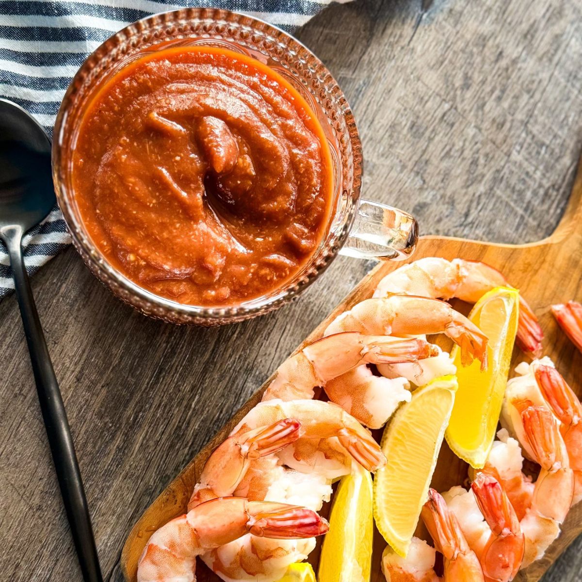 A glass bowl of cocktail sauce sits beside a wooden platter of cooked shrimp garnished with lemon wedges, all on a wooden surface with a striped cloth in the background.