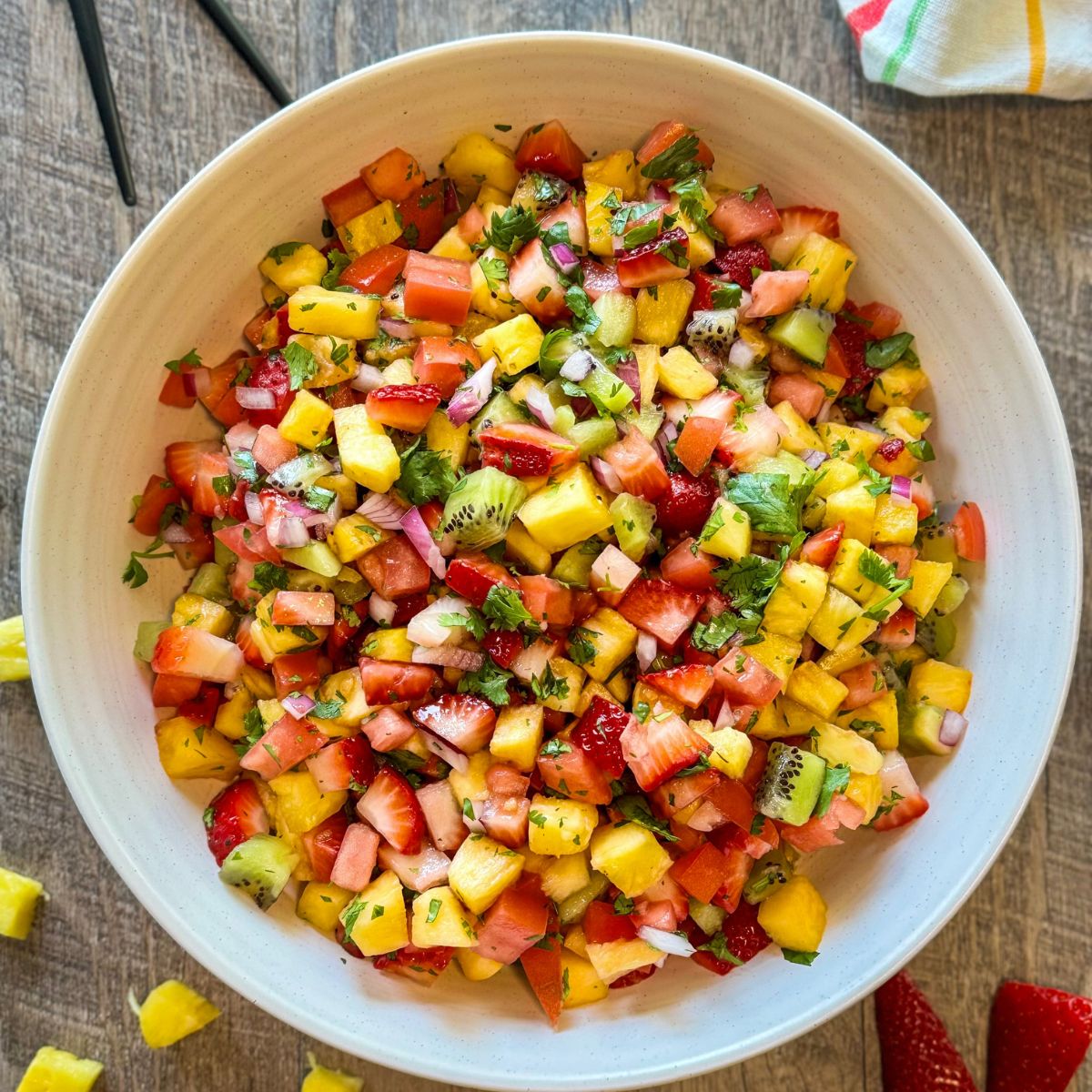 A large white bowl filled with a colorful fruit salsa made of chopped strawberries, kiwi, pineapple, mango, red onion, cilantro, and red bell pepper, placed on a wooden surface.