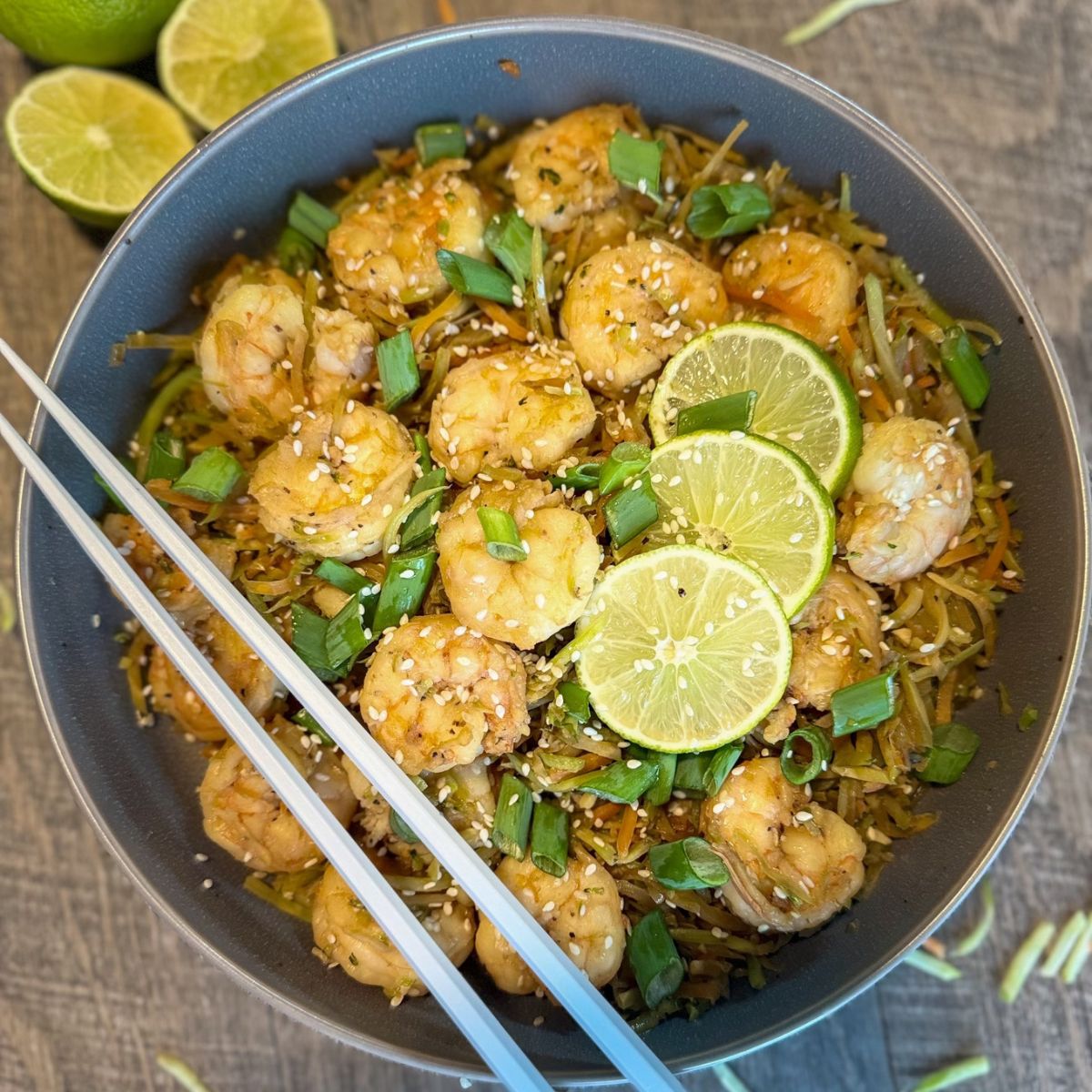 A bowl of shrimp stir fry with sliced green onions, sesame seeds, and lime slices on top, served with chopsticks on the side. Shredded vegetables and two lime halves are visible in the background.