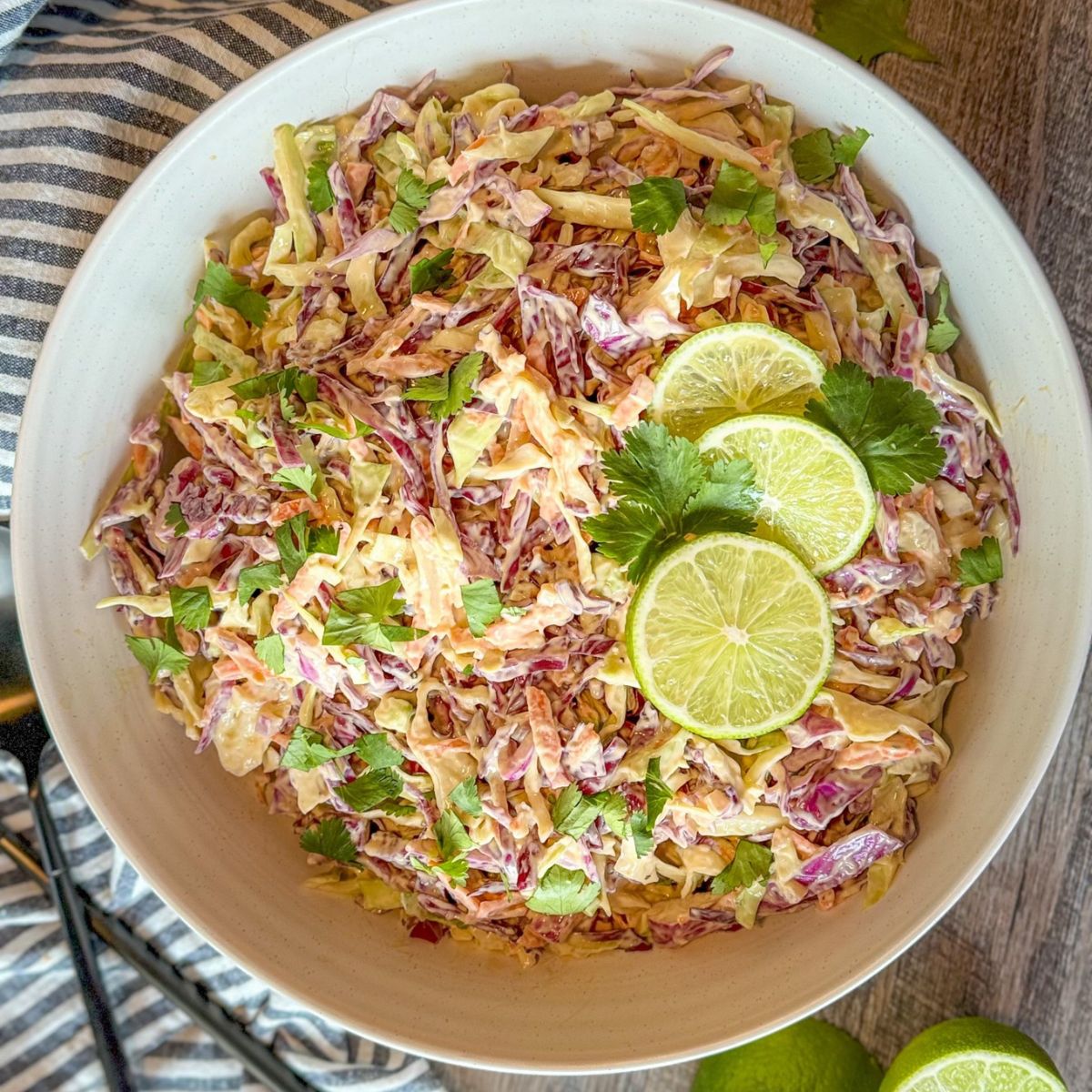 A bowl of colorful coleslaw made with shredded cabbage, carrots, and fresh cilantro, garnished with lime slices on top. The bowl sits on a striped cloth with limes and cilantro nearby.