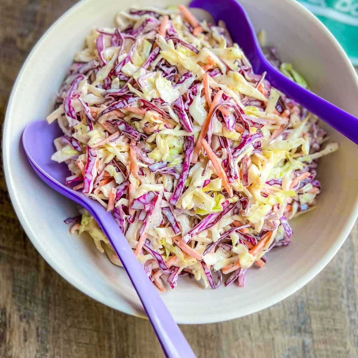 A bowl of colorful coleslaw made with shredded green and purple cabbage and carrots, served with two purple serving utensils on a wooden surface.