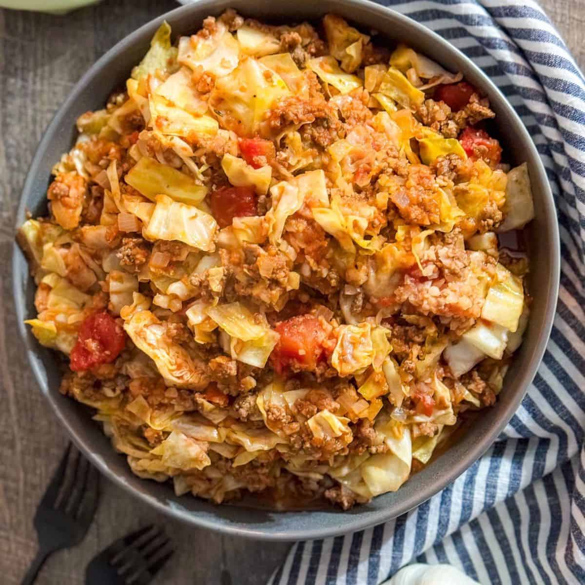 A bowl filled with unstuffed cabbage roll casserole, featuring chopped cabbage, ground meat, tomatoes, and rice, placed on a wooden table next to a striped cloth and black utensils.